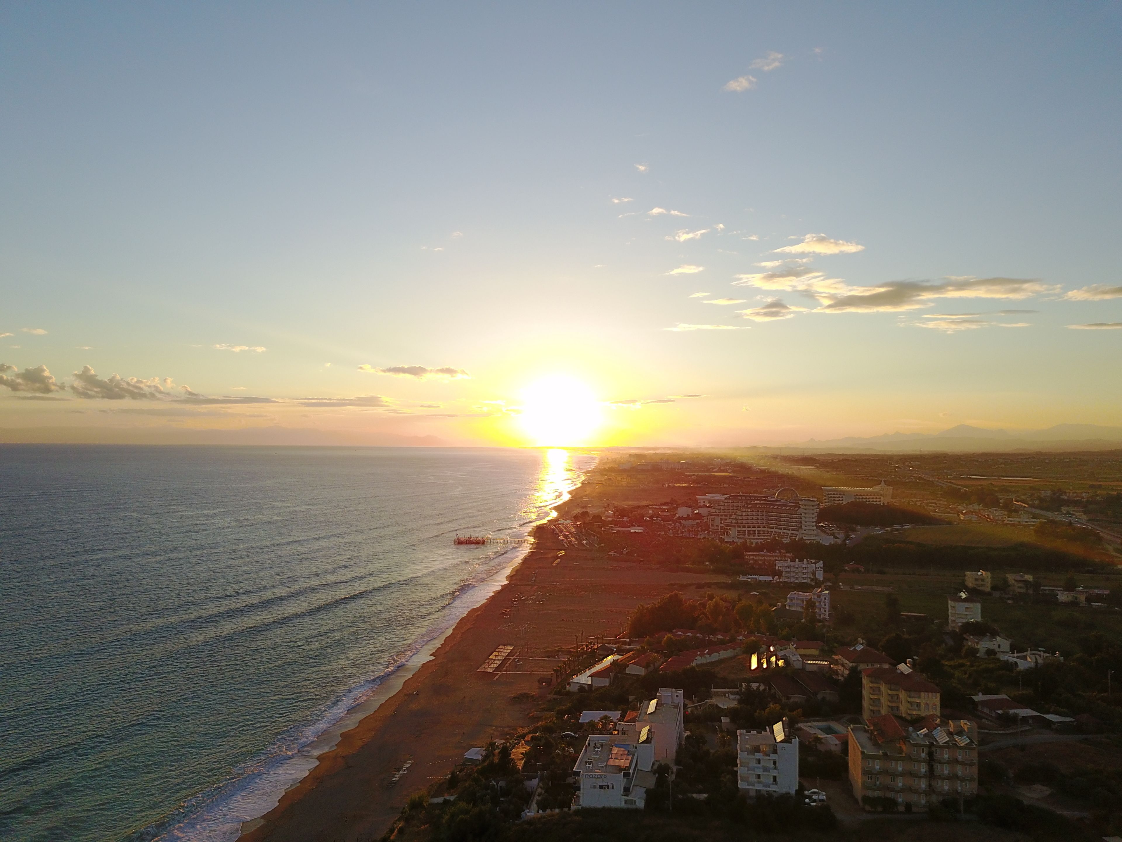 Luftaufnahme einer Küstenregion mit Strand, Gebäuden und einem klaren Himmel bei Sonnenuntergang.
