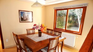 Une salle à manger avec une table en bois, des chaises, un vase de fleurs, un tableau encadré au mur et une fenêtre donnant sur un paysage verdoyant.