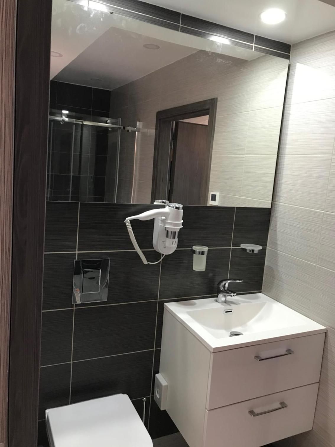 Un baño moderno con lavamanos blanco, paredes con azulejos oscuros, un espejo y una zona de ducha.