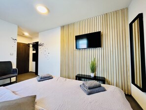 A modern hotel room with a large bed, a flat-screen TV mounted on the wall, a small table with a plant, and a seating area with a chair and a sofa. - Penzion Hornička (Cesky Krumlov)