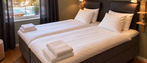 A double bed with white linens and two bedside lamps, a window with dark curtains, and a view of an outdoor pool area.