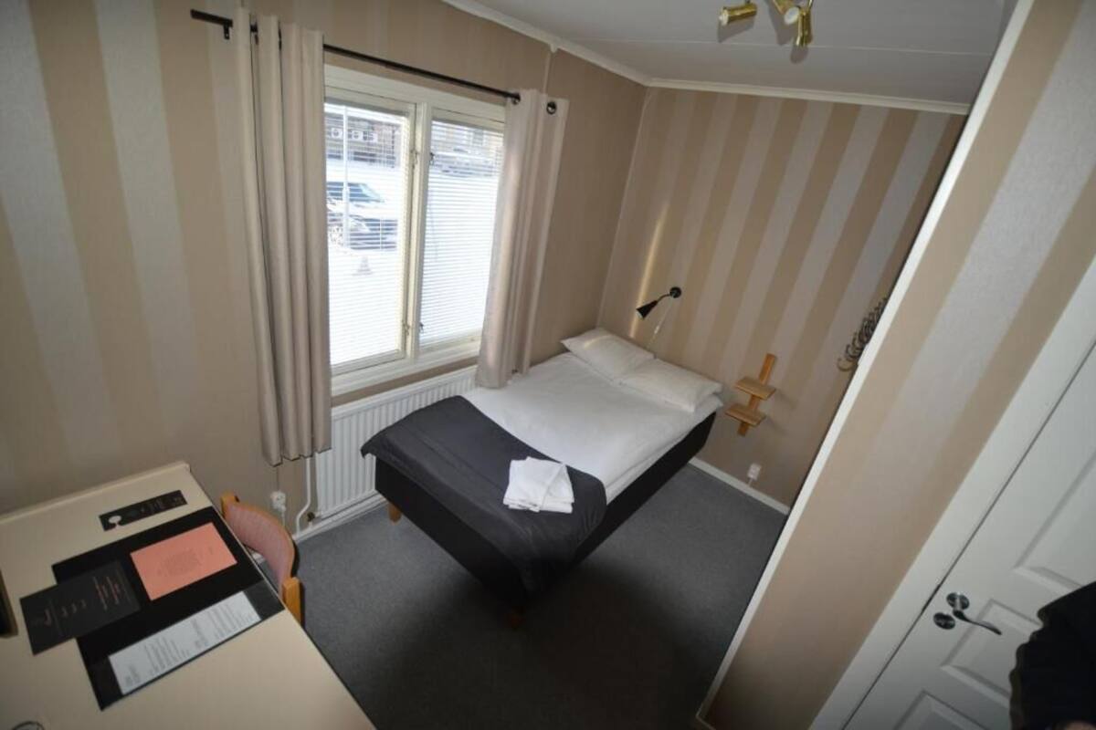 Ein kleines Hotelzimmer mit einem Einzelbett, einem Schreibtisch mit Unterlagen und einem Fenster mit Vorhängen.