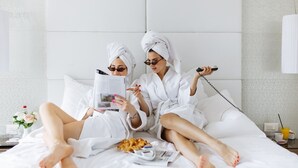 Twee personen in witte badjassen zitten op een bed met een tijdschrift, een telefoon en snacks.