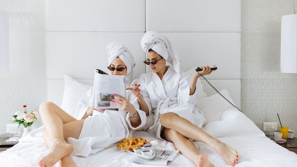 Zwei Personen in weißen Bademänteln sitzen auf einem Bett mit einer Zeitschrift, einem Telefon und Snacks.