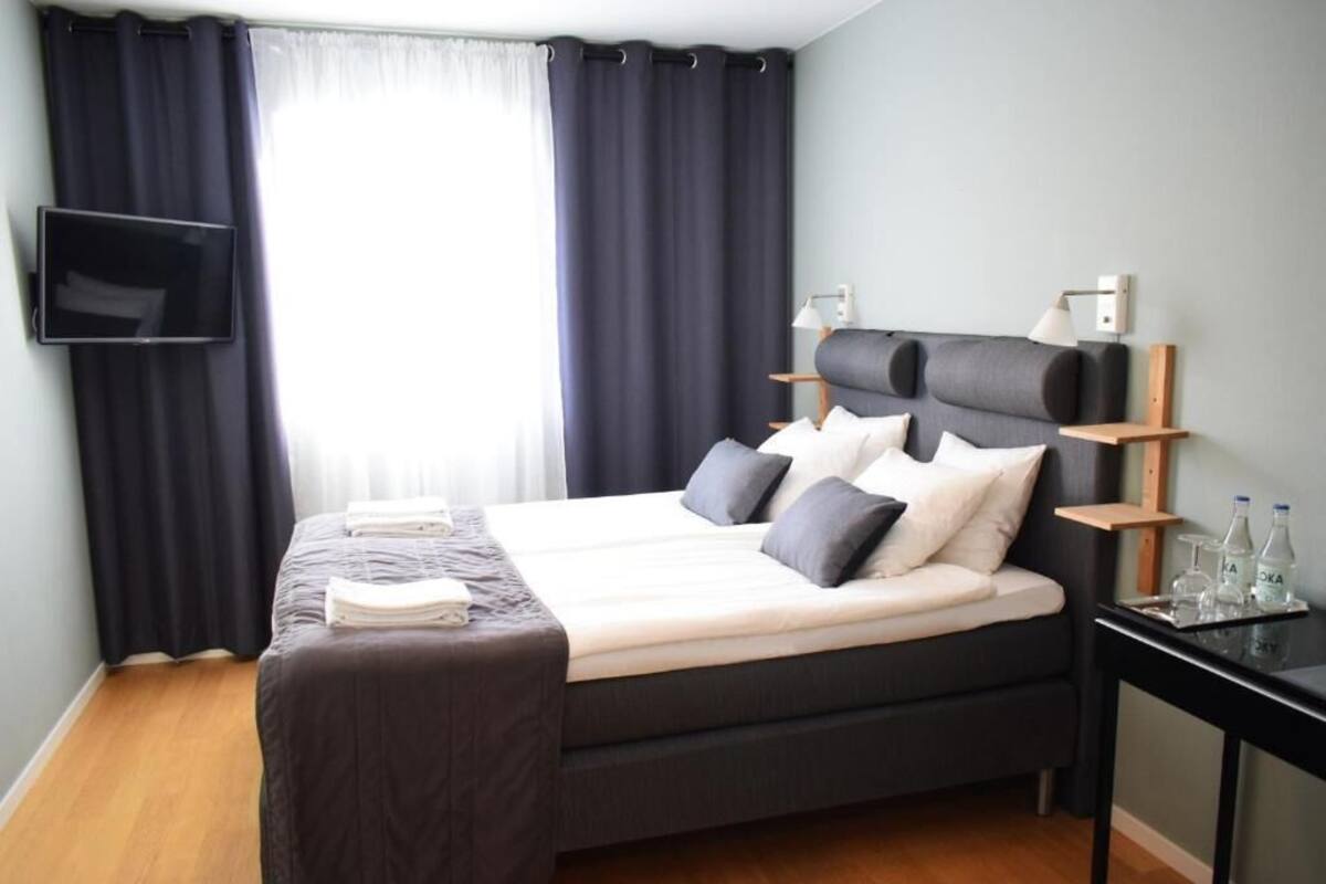 Ein modernes Hotelzimmer mit einem großen Bett, dunklen Vorhängen, einem flachbildfernseher an der Wand, einem Nachttisch mit Lampe und einer kleinen Ablagefläche mit Flaschen.