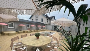 Eine Terrasse im Freien mit einem runden Holztisch und Stühlen, umgeben von gepolsterten Sitzgelegenheiten und Sonnenschirmen.