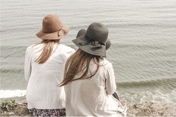 Dos personas de pie junto al mar, usando sombreros y ropa informal.