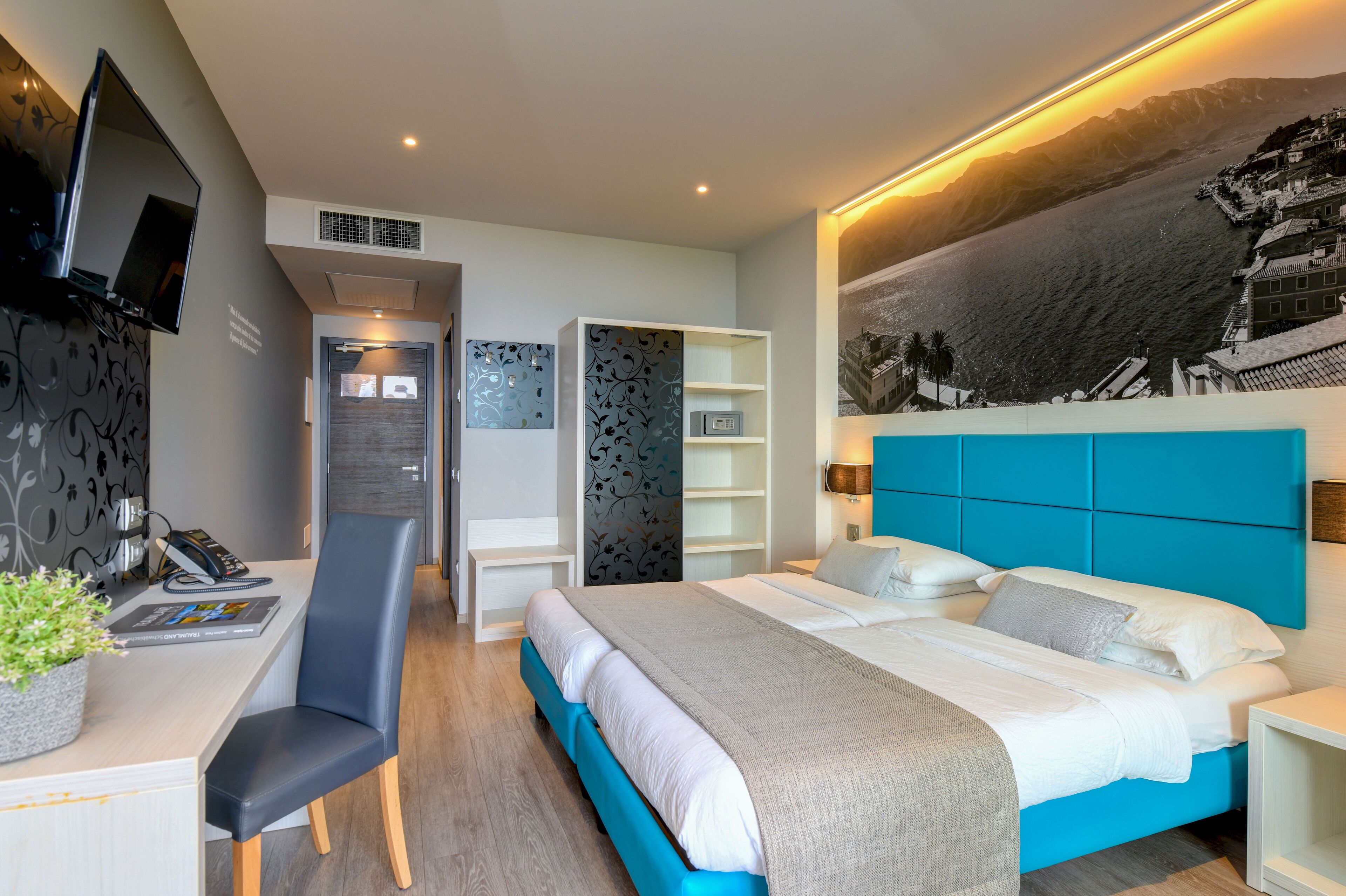 A modern hotel room with a large bed, a desk with a chair, a flat-screen TV, and a view of the sea.