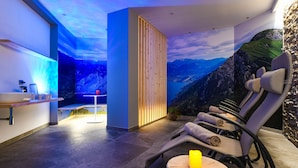 Ein moderner Wellnessraum mit Blick auf die Berge, einem Holzkabinett und einer Entspannungszone mit Sesseln und einem kleinen Tisch.
