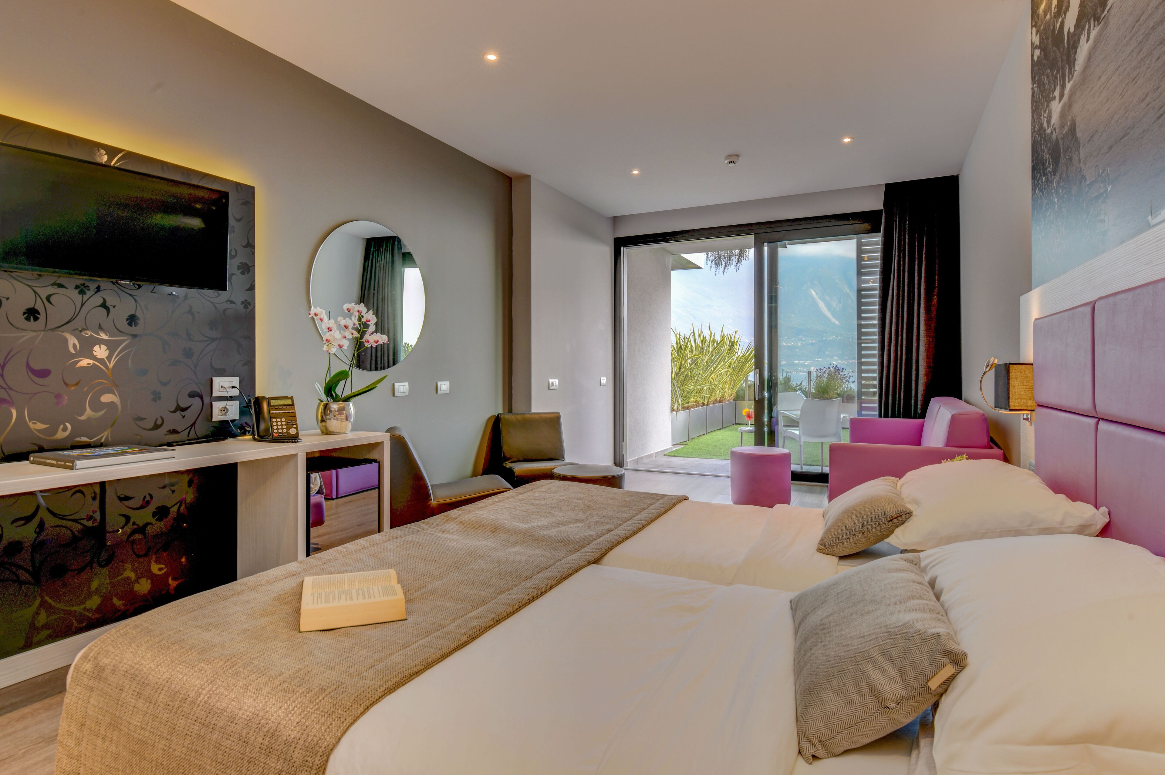 A modern hotel room with a large bed, a desk with a flat-screen TV, a chair, a small table, and a view of the outdoors.