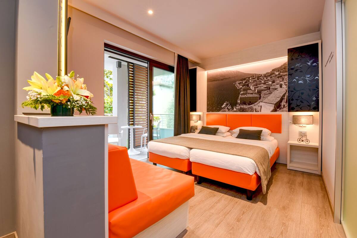 Una habitación de hotel moderna con una cama grande, dos mesitas de noche, un sofá y un balcón con vistas a un pueblo.