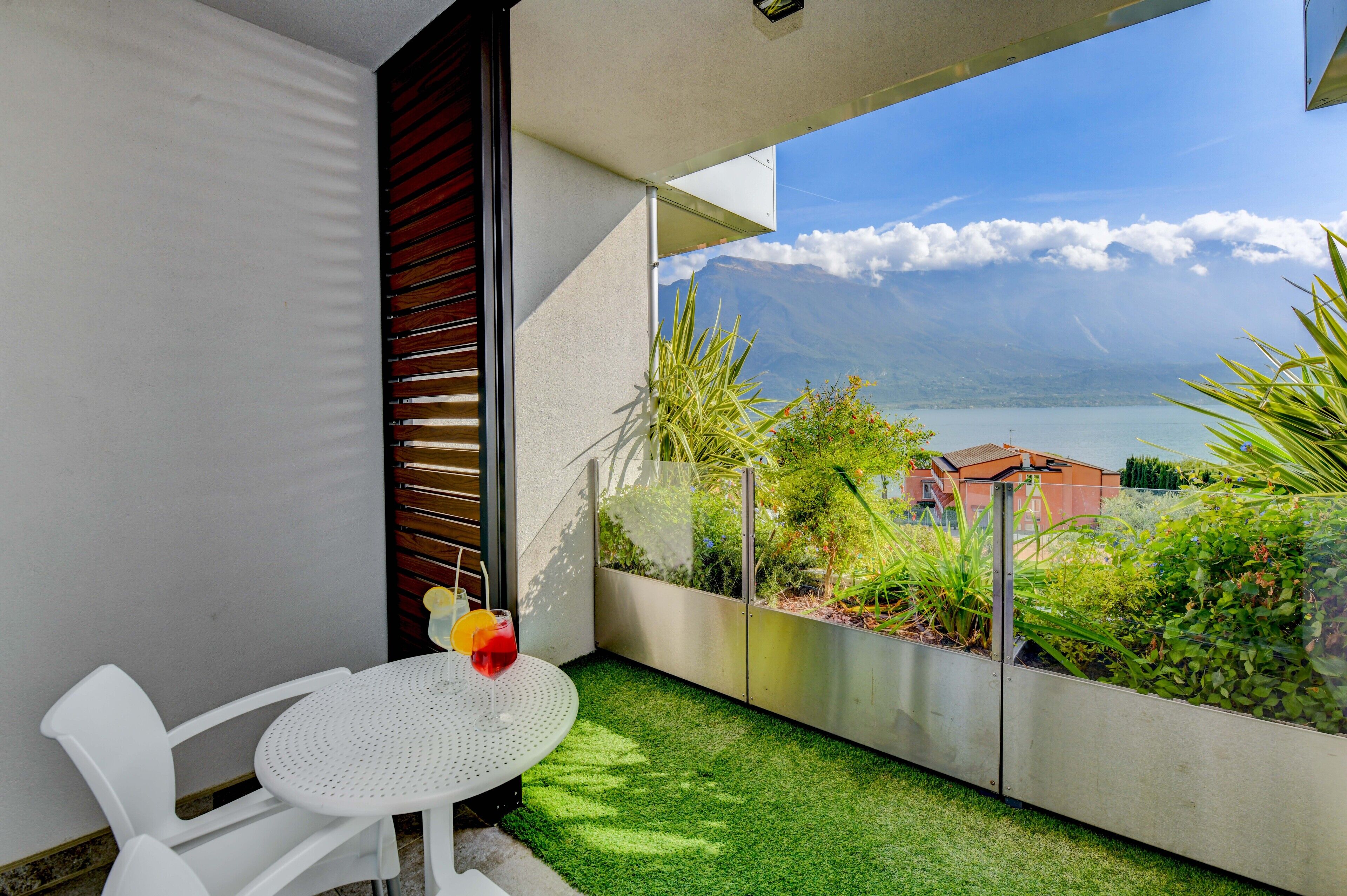 A balcony with a white chair, a small round table, and a drink. It overlooks a garden with various plants and a view of a lake and mountains in the distance.