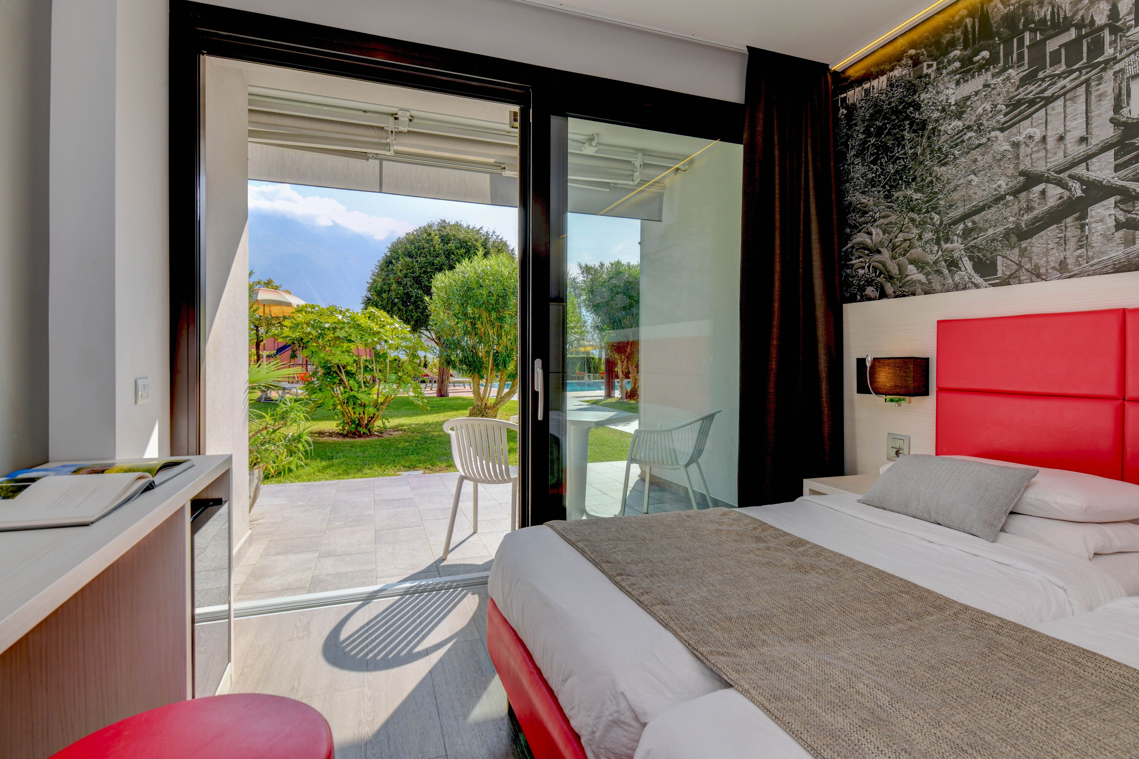 Een moderne hotelkamer met een groot bed, een bureau en een glazen deur die leidt naar een buitenruimte met een zwembad en groen.
