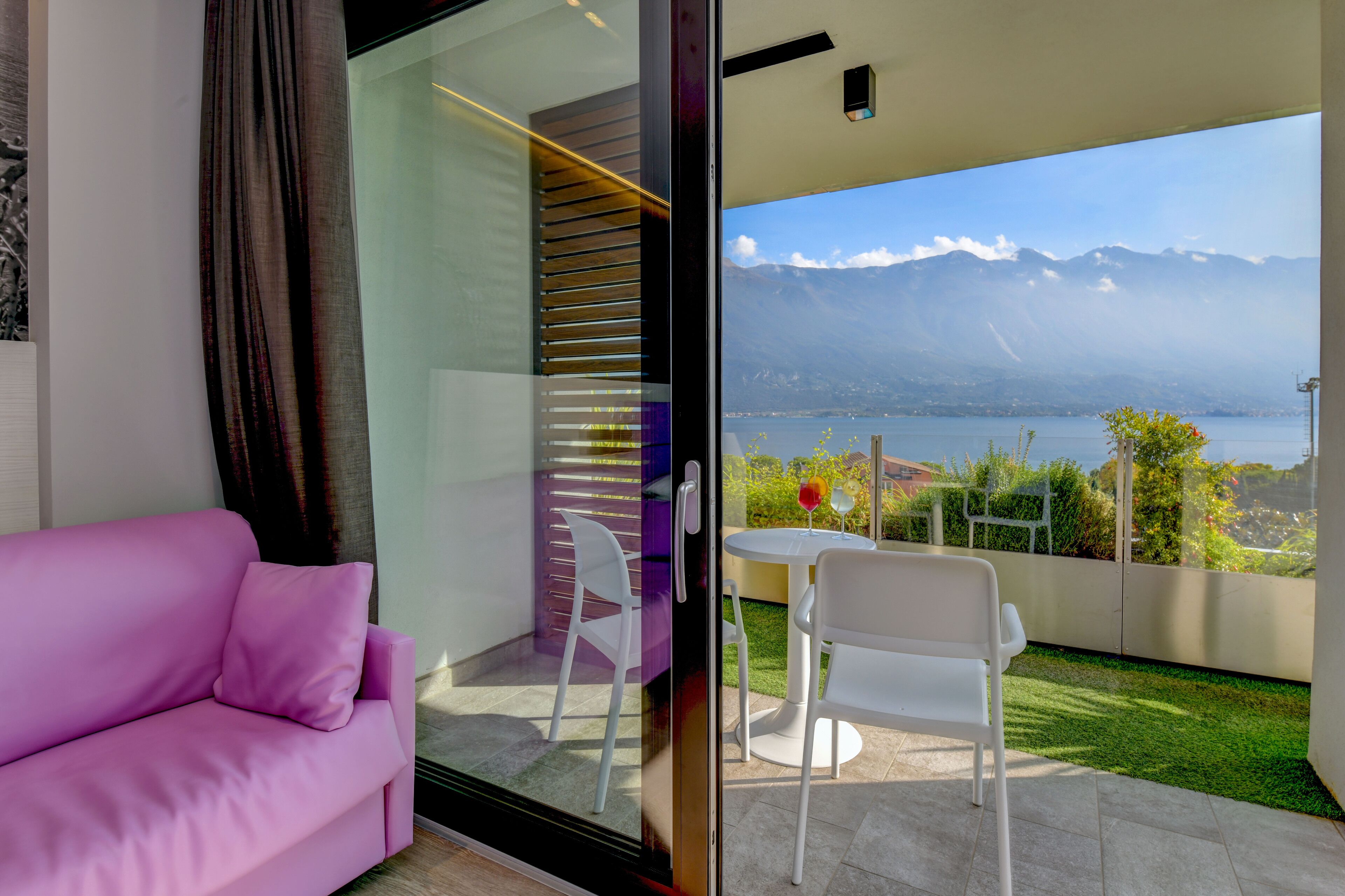 Un balcón moderno con puerta de cristal, un sofá rosa, sillas blancas y una mesa.