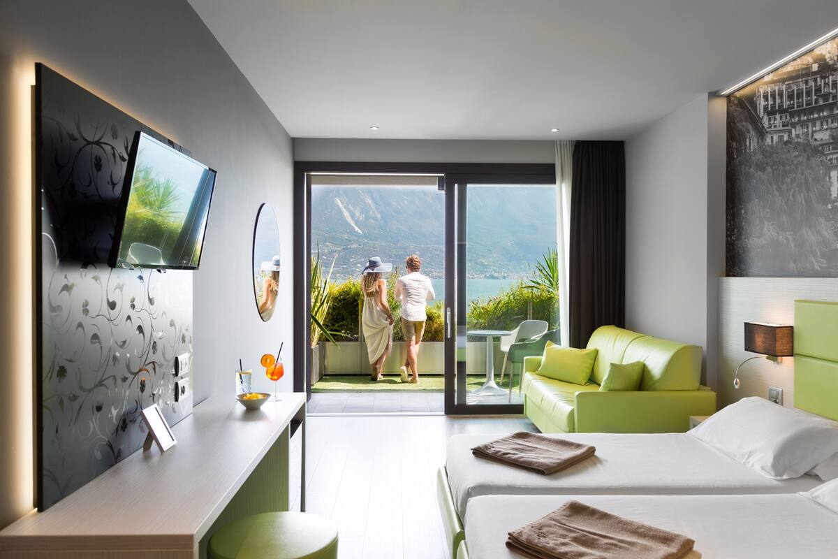 Una habitación de hotel moderna con una cama grande, un sofá verde, un escritorio con una computadora portátil y un balcón con vistas a las montañas.
