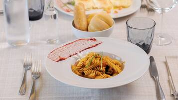 A dining table with a plate of pasta, a glass of water, and a bowl of bread rolls.