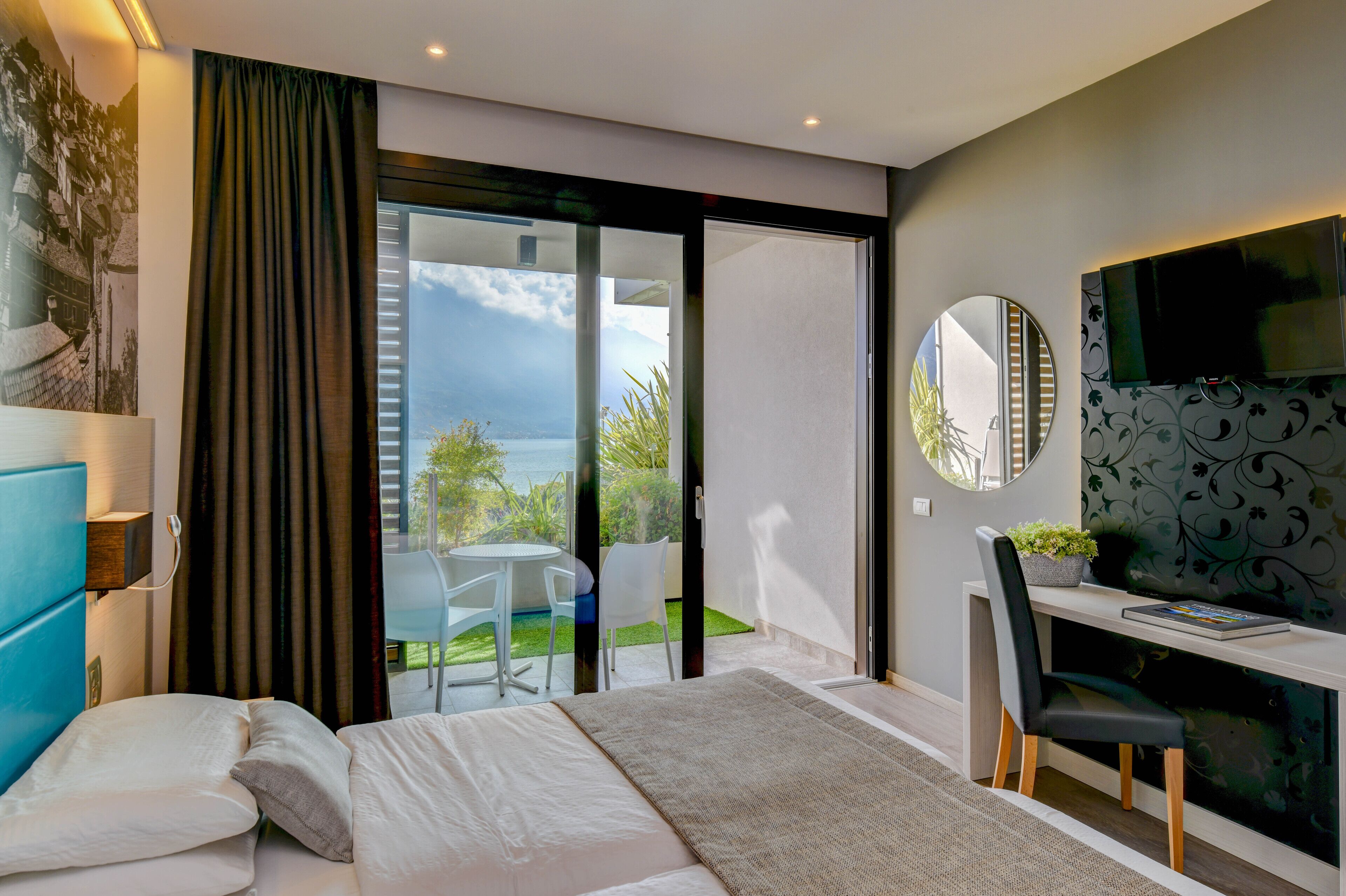 A modern bedroom with a bed, a desk, and a TV. There is a sliding glass door leading to a balcony with a view of the sky and greenery.