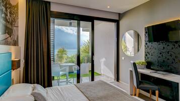 A modern bedroom with a bed, a desk, and a TV. There is a sliding glass door leading to a balcony with a view of the sky and greenery.