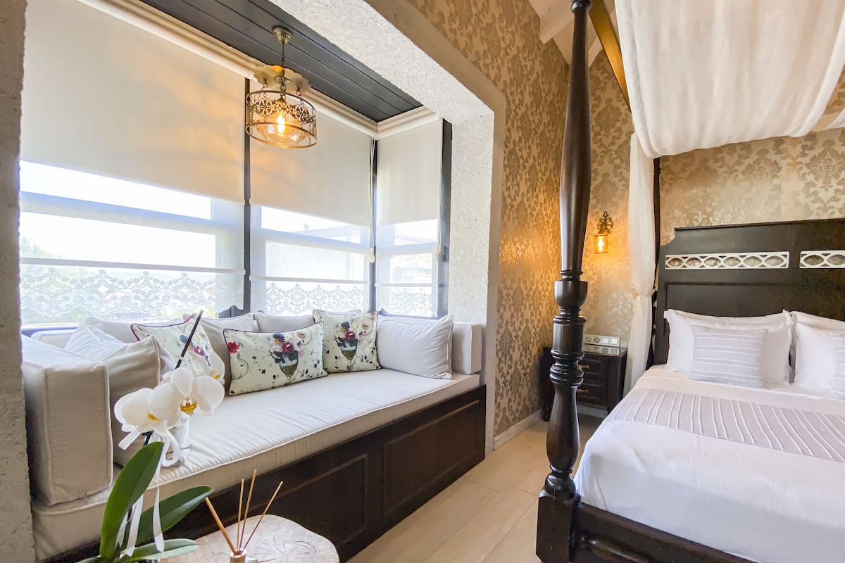 A bedroom with a four-poster bed, a sofa bed with decorative pillows, a nightstand, and a large window with a view.