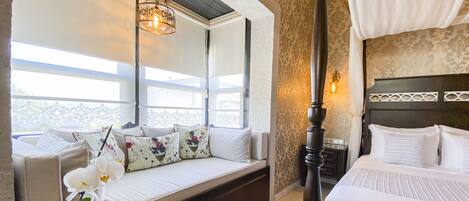 A bedroom with a four-poster bed, a sofa bed with decorative pillows, a nightstand, and a large window with a view.