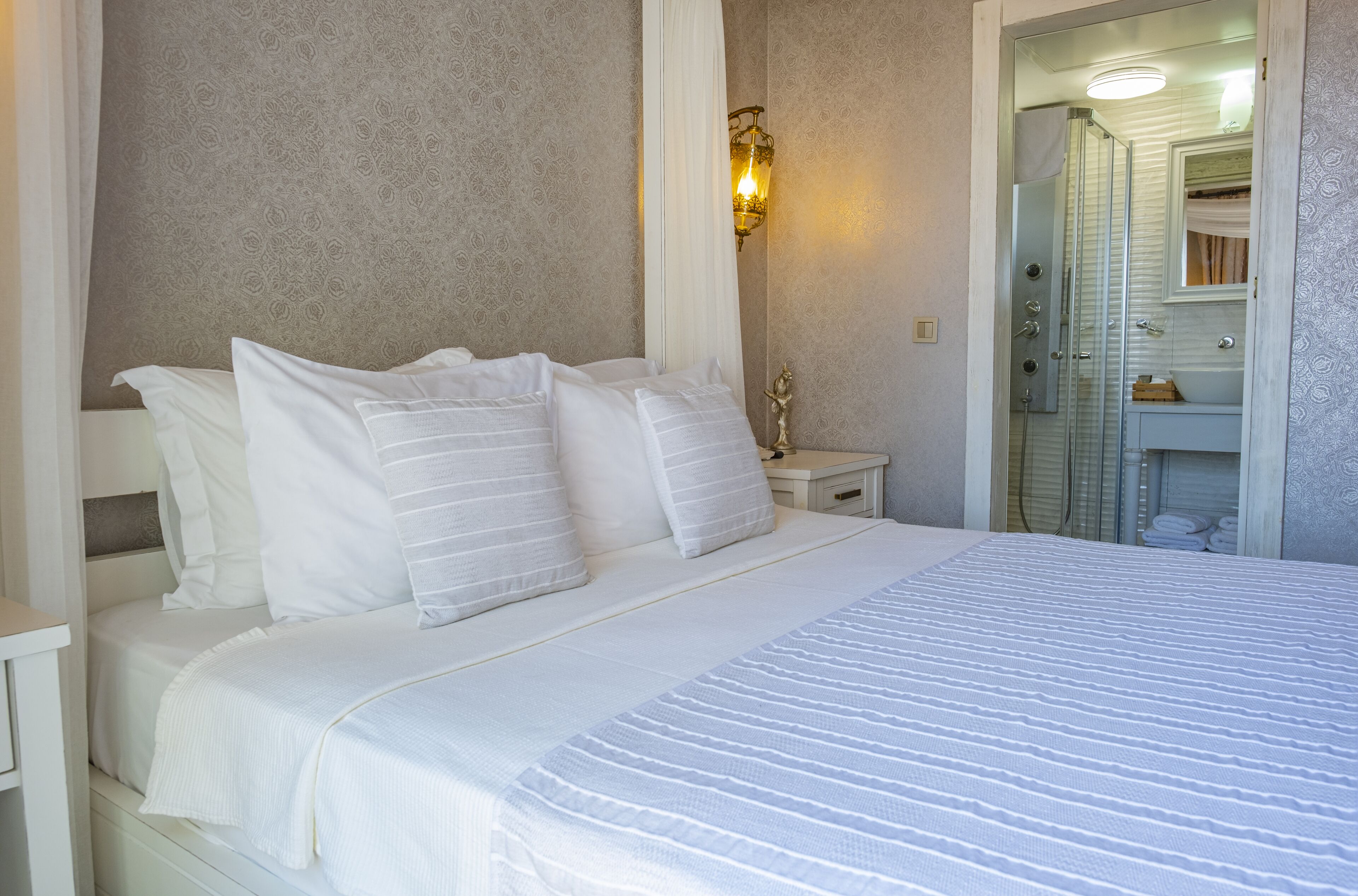 A neatly made bed with white linens and pillows, a bedside table with a lamp, and a bathroom visible through an open door.