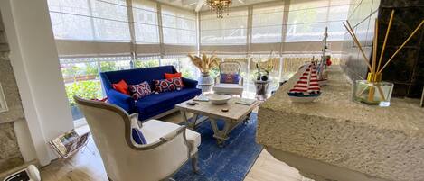 A sunlit room with a blue sofa, white armchairs, a dining table, and a stone fireplace.