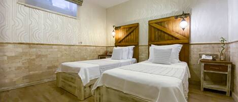 A room with two beds, a wooden floor, a window with a patterned curtain, and a wall-mounted light fixture.