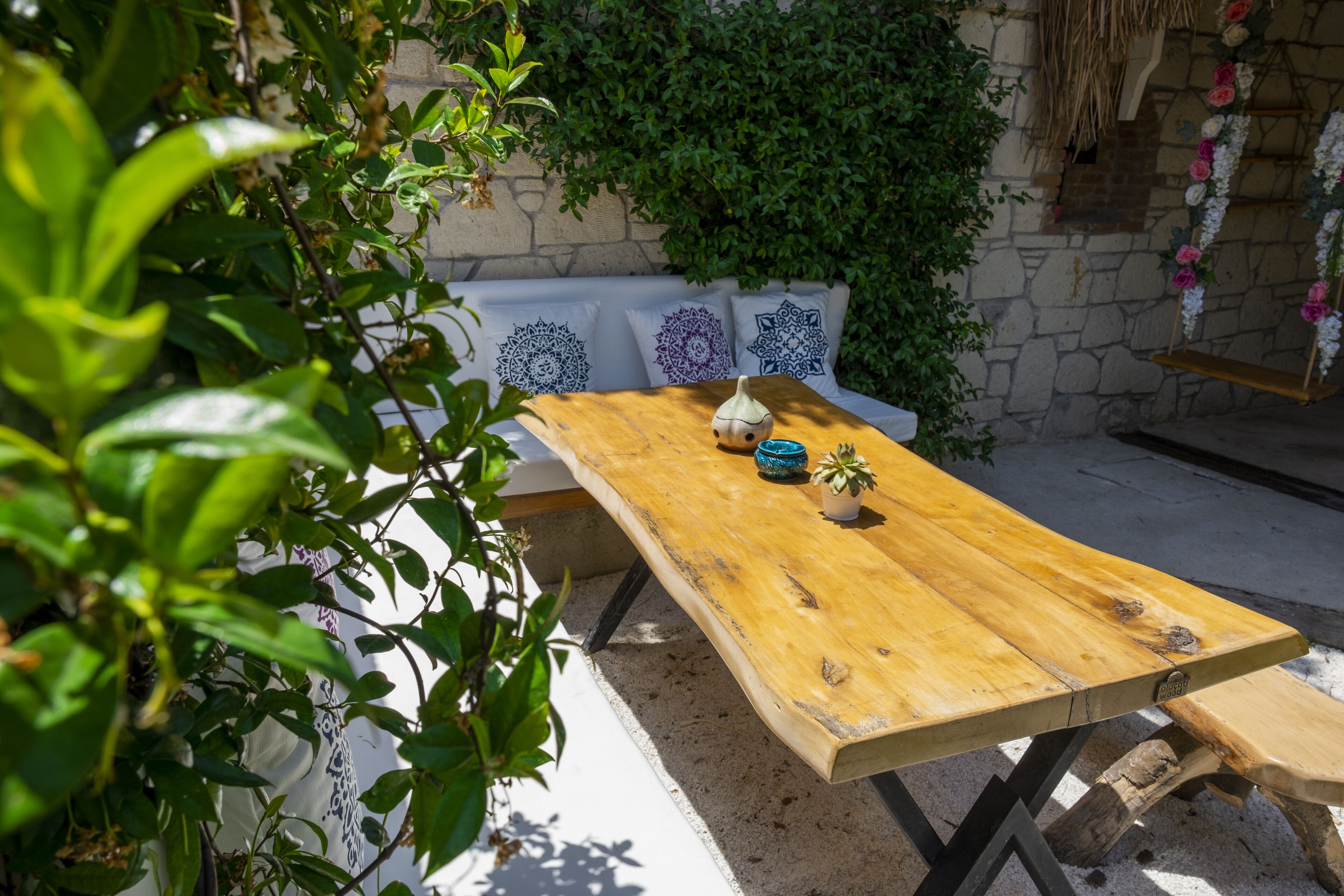 A wooden table with a cushion bench, surrounded by greenery and a stone wall.
