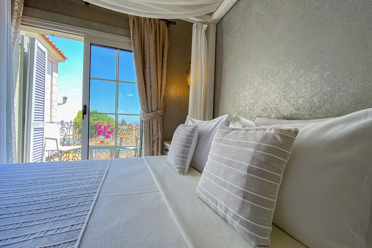 A bedroom with a large bed, white bedding, and a view of a balcony with outdoor furniture and plants.