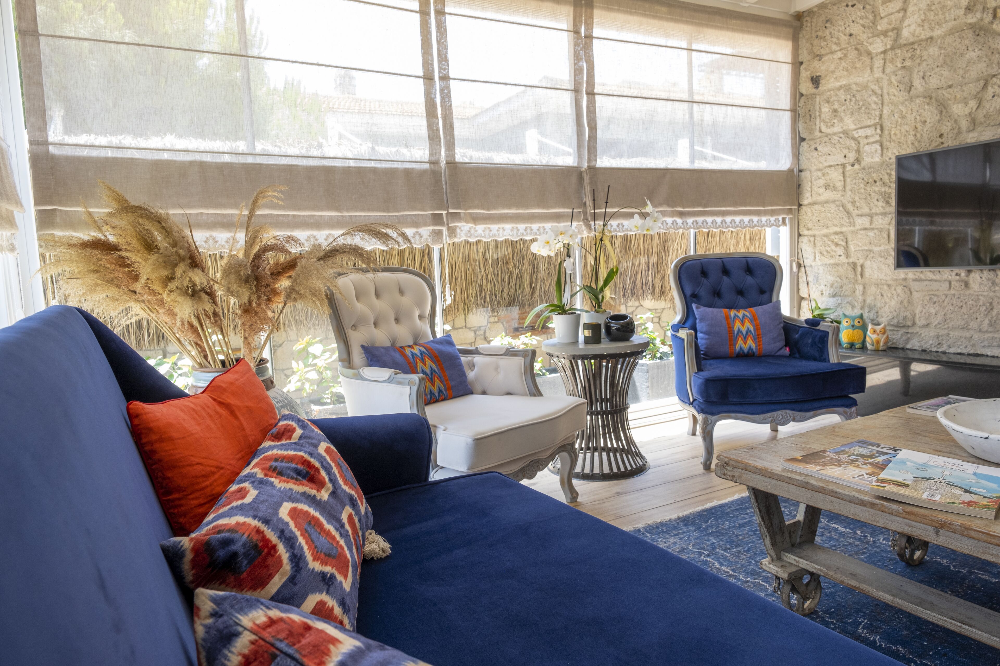A cozy living room with a blue sofa, a white armchair, and a blue armchair with patterned cushions. There is a coffee table with magazines and a decorative bowl. The room has a stone wall and a large window with a view of the outdoors.