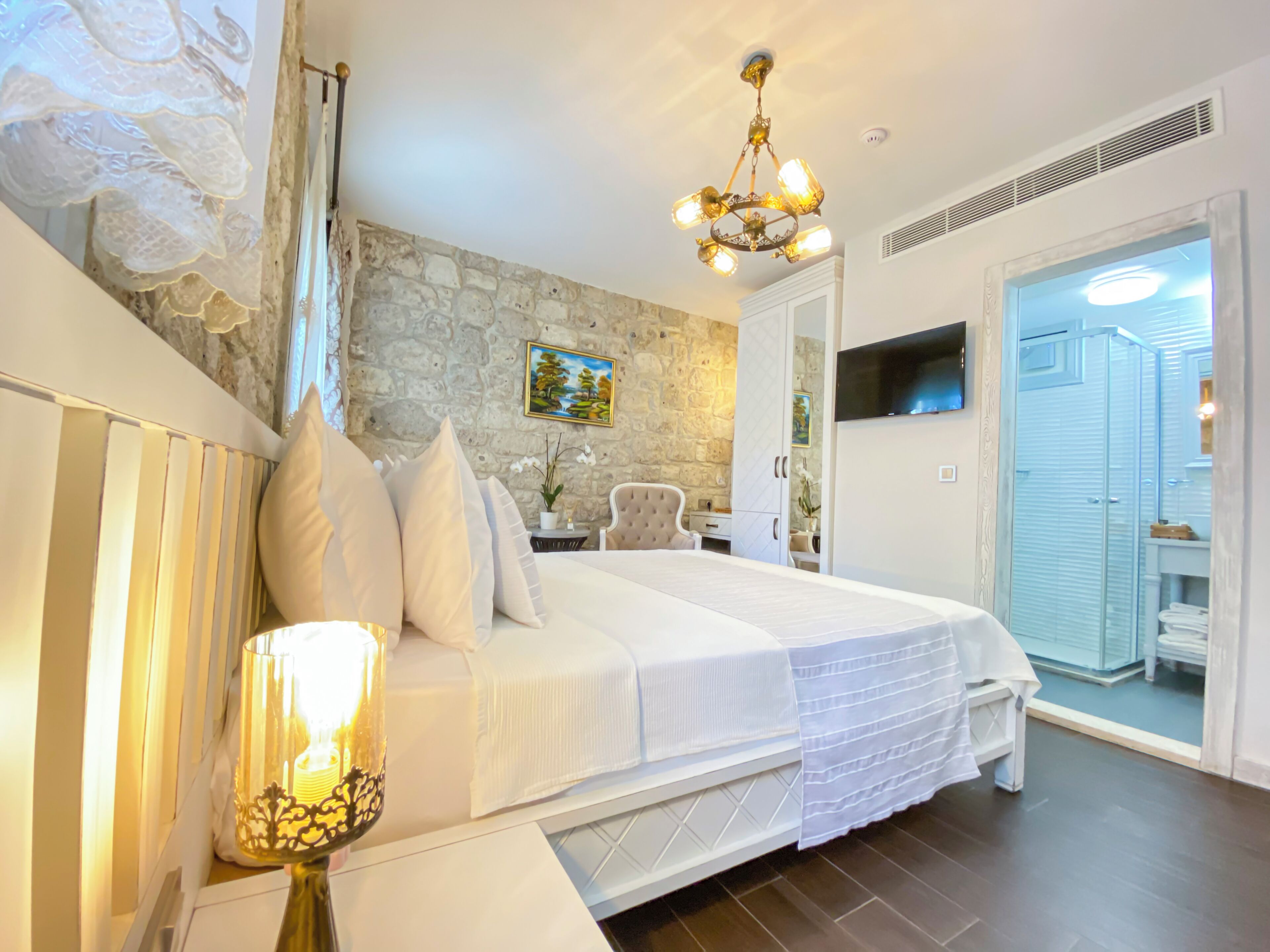 A bedroom with a stone wall, a bed with white bedding, a chandelier, a TV, and a bathroom visible through an open door.