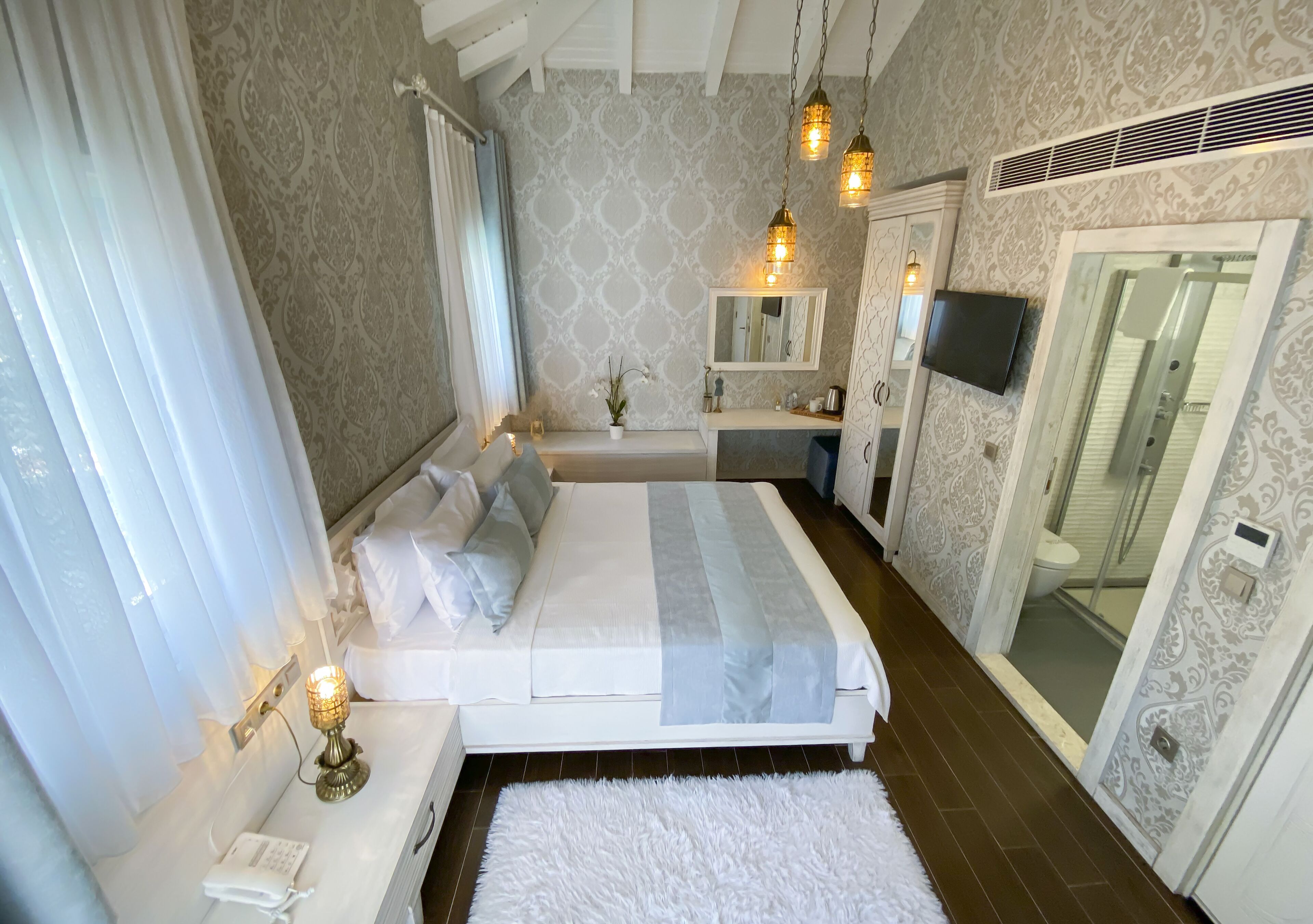 A bedroom with a bed, bedside table, lamp, and a bathroom with a shower, toilet, and mirror.