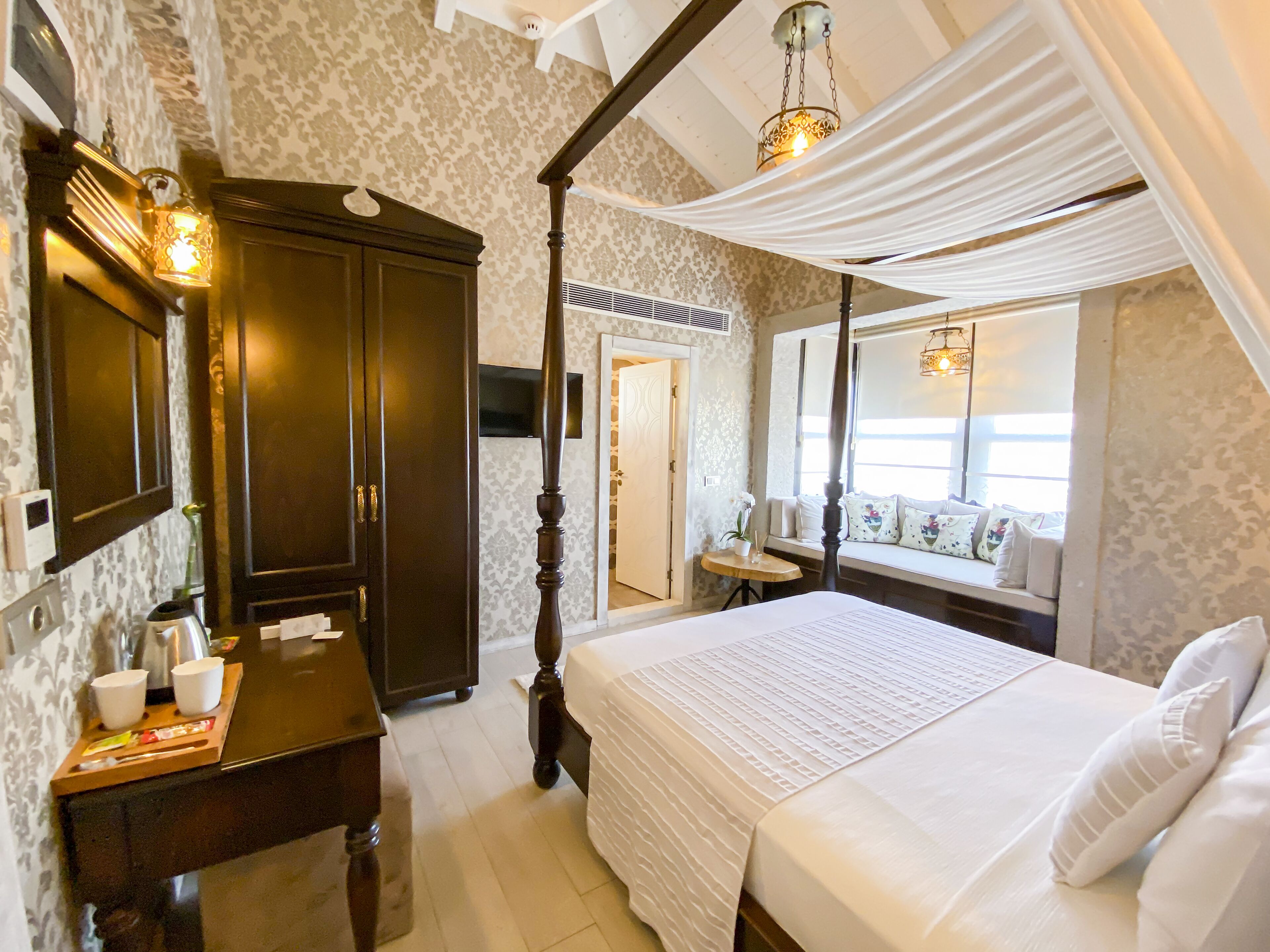 A bedroom with a four-poster bed, a wooden wardrobe, a small table with cups and a tray, and a window seat with cushions.