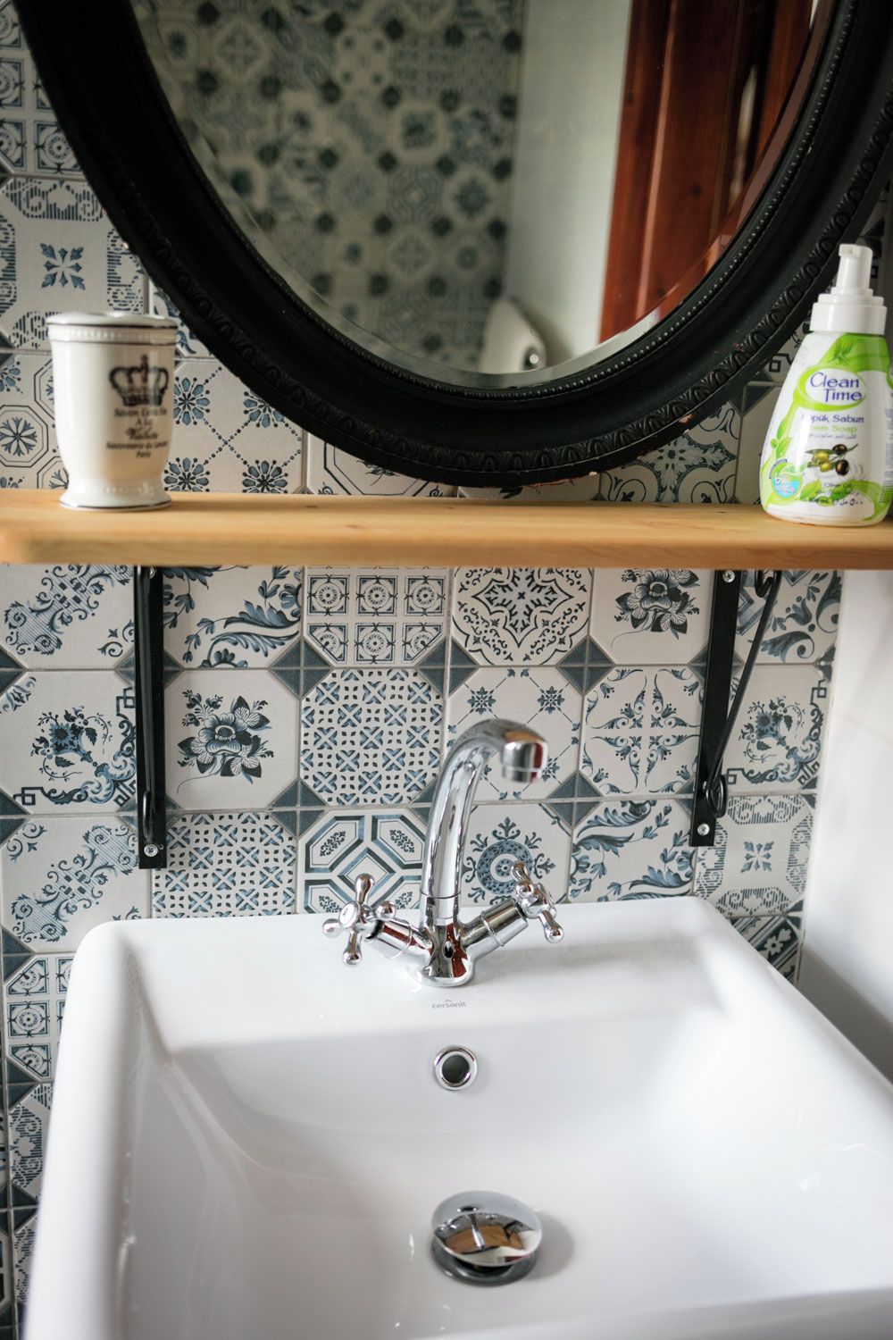 A bathroom with a white sink, a black faucet, a mirror above the sink, and a shelf with a cup and a bottle of cleaning product.