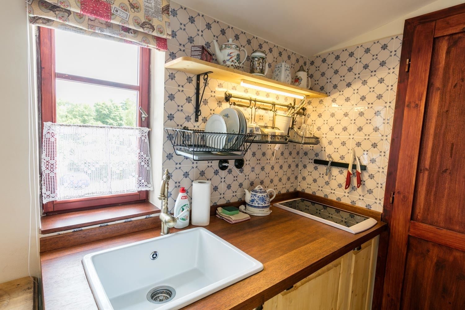 A kitchen with a wooden countertop, a white sink, a window with lace curtains, and a shelf with dishes and a kettle.