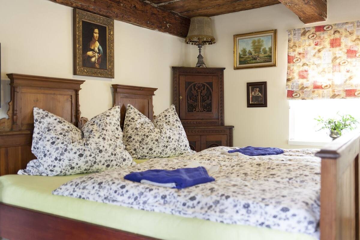 Quarto com cama, cabeceira de madeira, almofadas estampadas com padrão floral, um armário de madeira e uma janela com cortina.