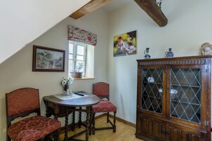 Quarto com mesa de madeira, duas cadeiras, um armário, janela com cortina e duas pinturas na parede.