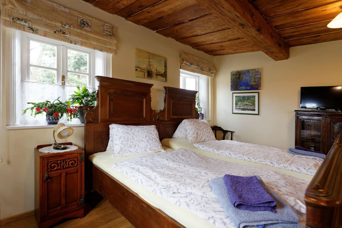Quarto com duas camas, cabeceiras de madeira, mesa de cabeceira, luminária, televisão e janela com cortinas.