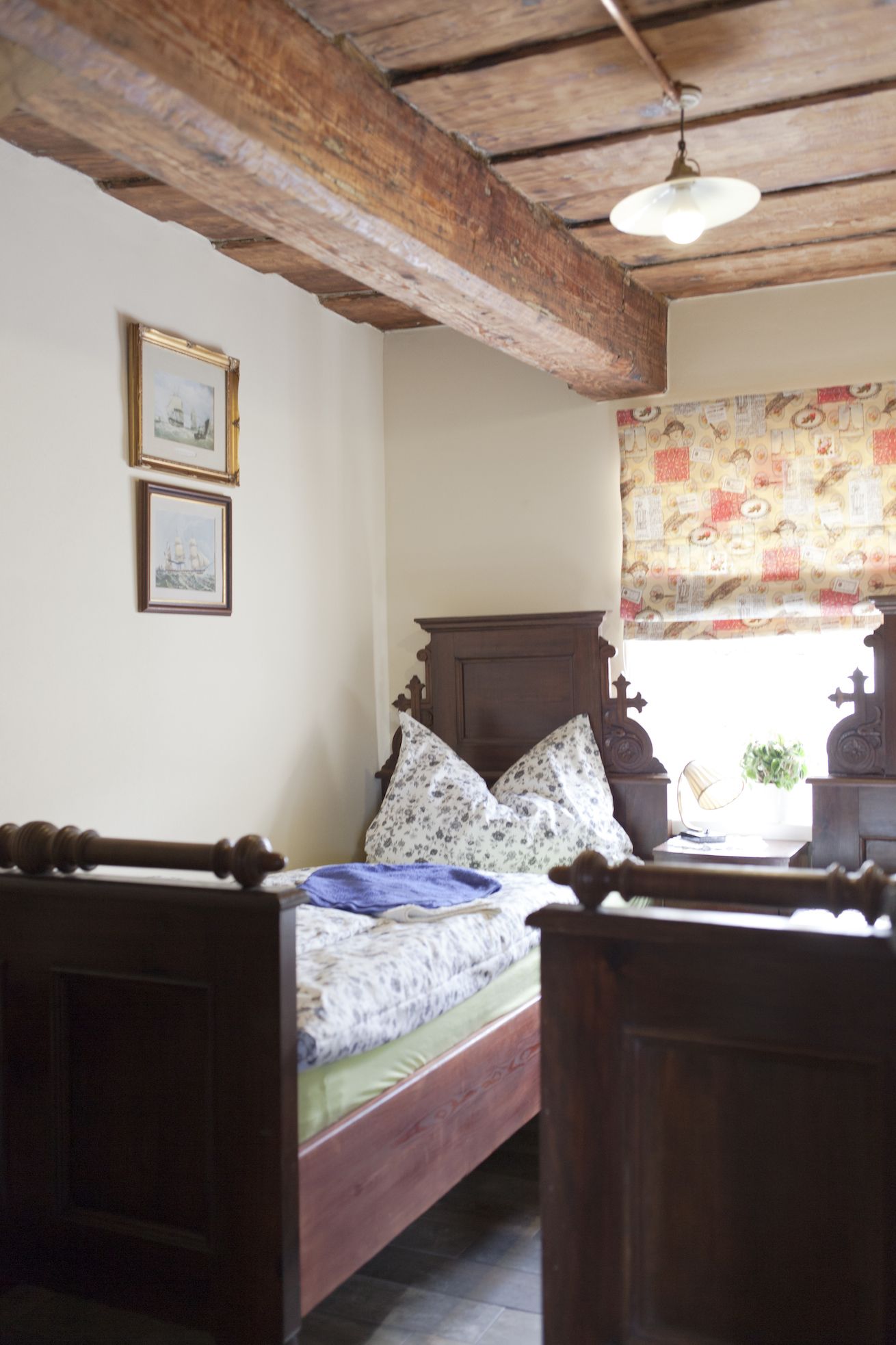 Une chambre à coucher avec un lit en bois, une tête de lit en bois, une fenêtre ornée d’un rideau à motifs et deux tableaux encadrés accrochés au mur.