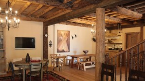 Een rustiek interieur met zichtbare houten balken, een kroonluchter, een eethoek met houten meubels, en een keuken in de achtergrond.