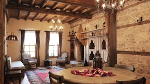 Een kamer in traditionele stijl met een houten plafond, stenen muren, een ronde tafel met stoelen, een kroonluchter en een open haard.