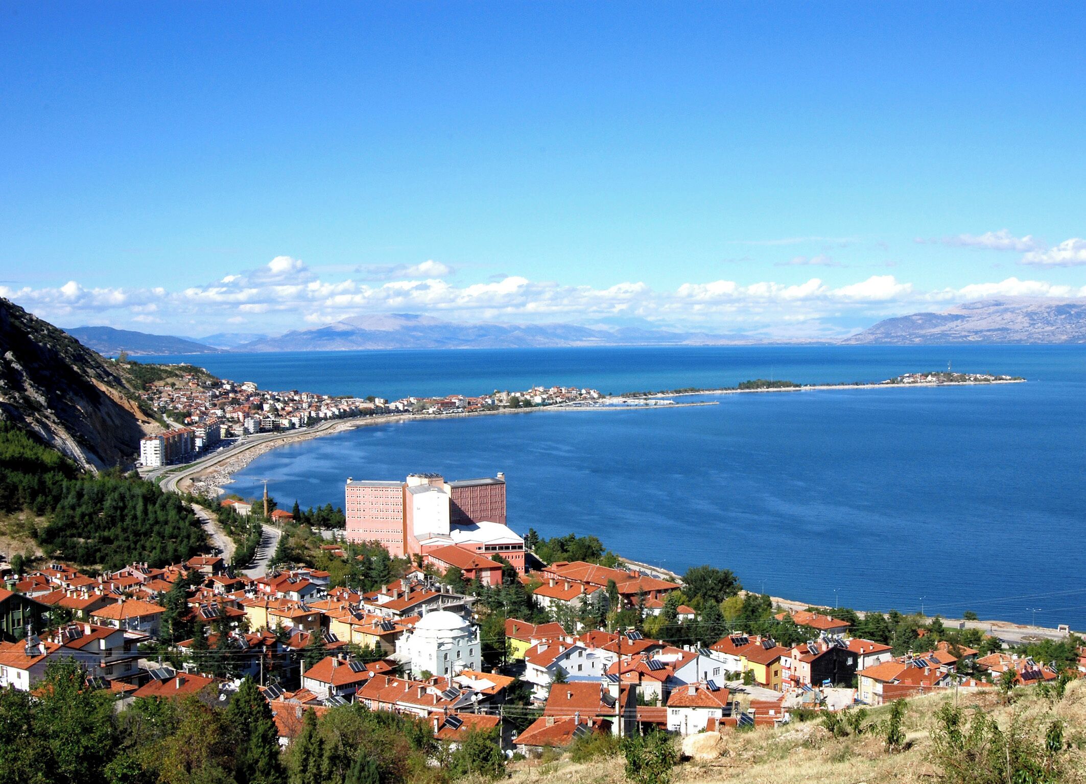 Une ville côtière aux bâtiments aux toits rouges, avec une église blanche imposante, et une vue sur la mer et les montagnes lointaines.