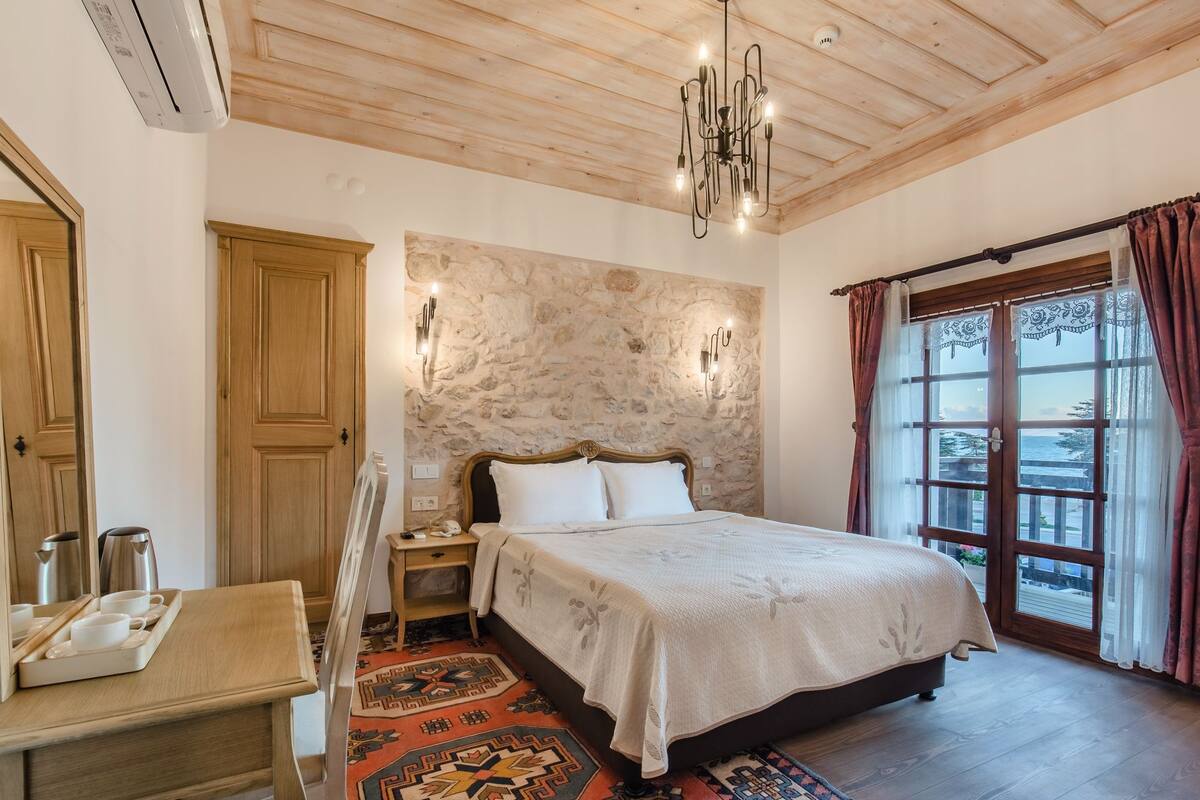 A bedroom with a bed, wooden ceiling, stone wall, and a view through a window with curtains.
