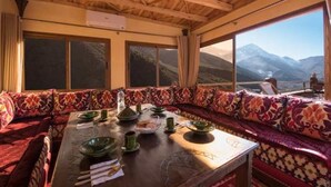 Un coin repas avec une table en bois dressée pour le repas, de grandes fenêtres offrant une vue sur les montagnes et un canapé à motifs.