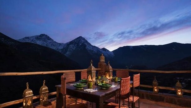 Une table à manger dressée avec des assiettes et des verres, entourée de chaises, avec une vue sur les montagnes en arrière-plan.