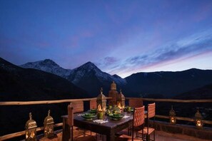A dining table set with plates and glasses, surrounded by chairs, with mountain views in the background. - Kasbah Imlil (Asni)