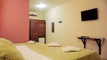 Quarto de hotel com uma cama, televisão na parede, uma mesinha e uma janela com persianas.