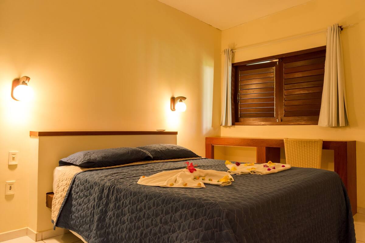 Un lit bien fait, recouvert d’une couette bleue, avec une tête de lit en bois et une fenêtre dotée de volets en bois.