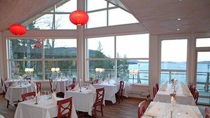 Un restaurant avec de nombreuses tables dressées pour le dîner, de grandes fenêtres offrant une vue sur l’eau et des lanternes rouges suspendues au plafond.