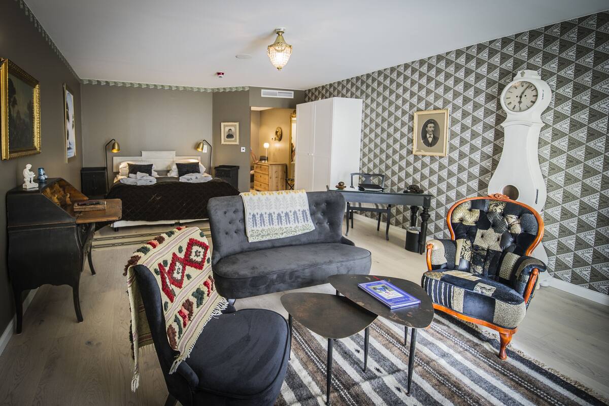 A well-furnished room with a bed, a sofa, a chair, a small table, and a clock on the wall.